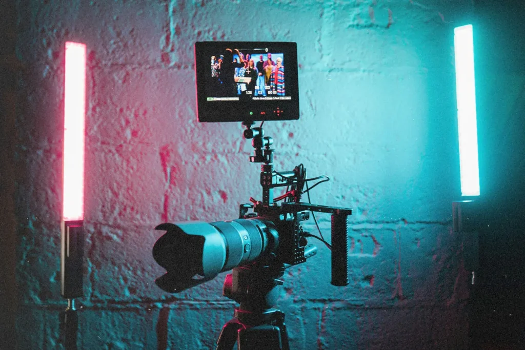 A professional video camera on a tripod is set up between two vertical LED lights glowing red and blue against a textured wall