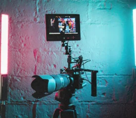 A professional video camera on a tripod is set up between two vertical LED lights glowing red and blue against a textured wall