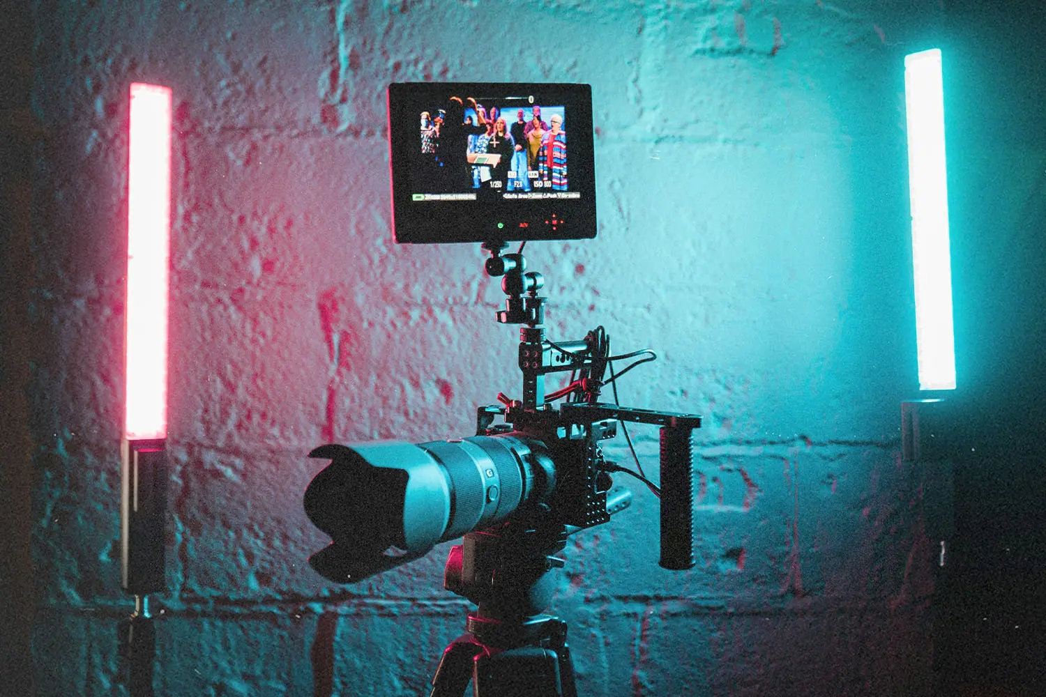 A professional video camera on a tripod is set up between two vertical LED lights glowing red and blue against a textured wall