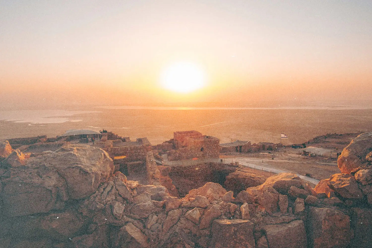 Golden sunrise over ancient stone ruins and a vast desert landscape with distant water reflecting the light