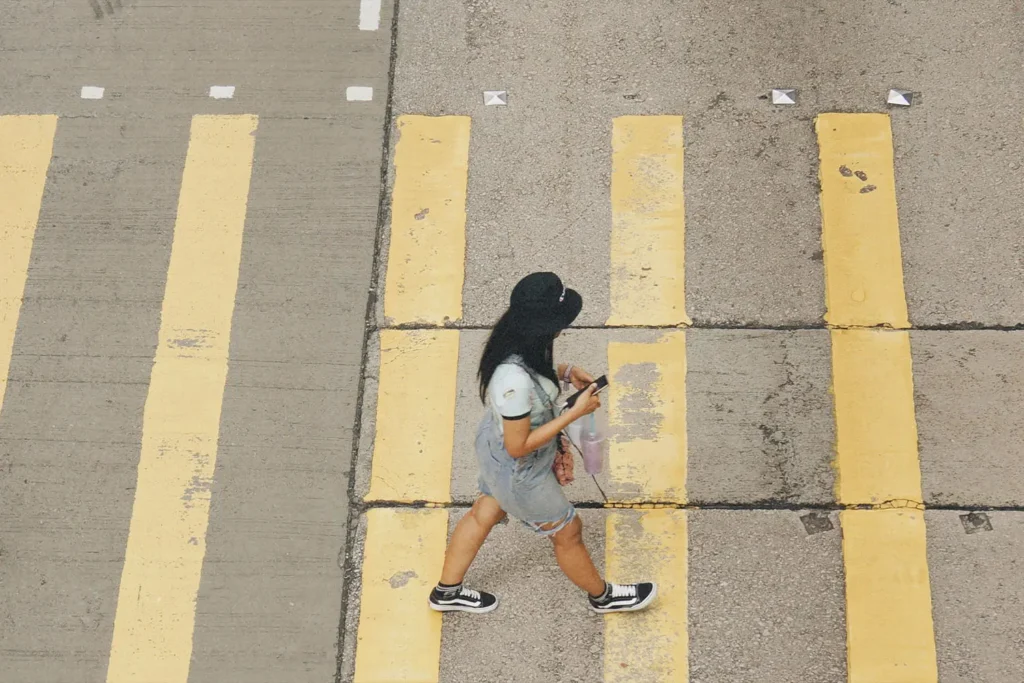 Person walking across a street with wide yellow crosswalk lines while holding a drink and looking at their phone