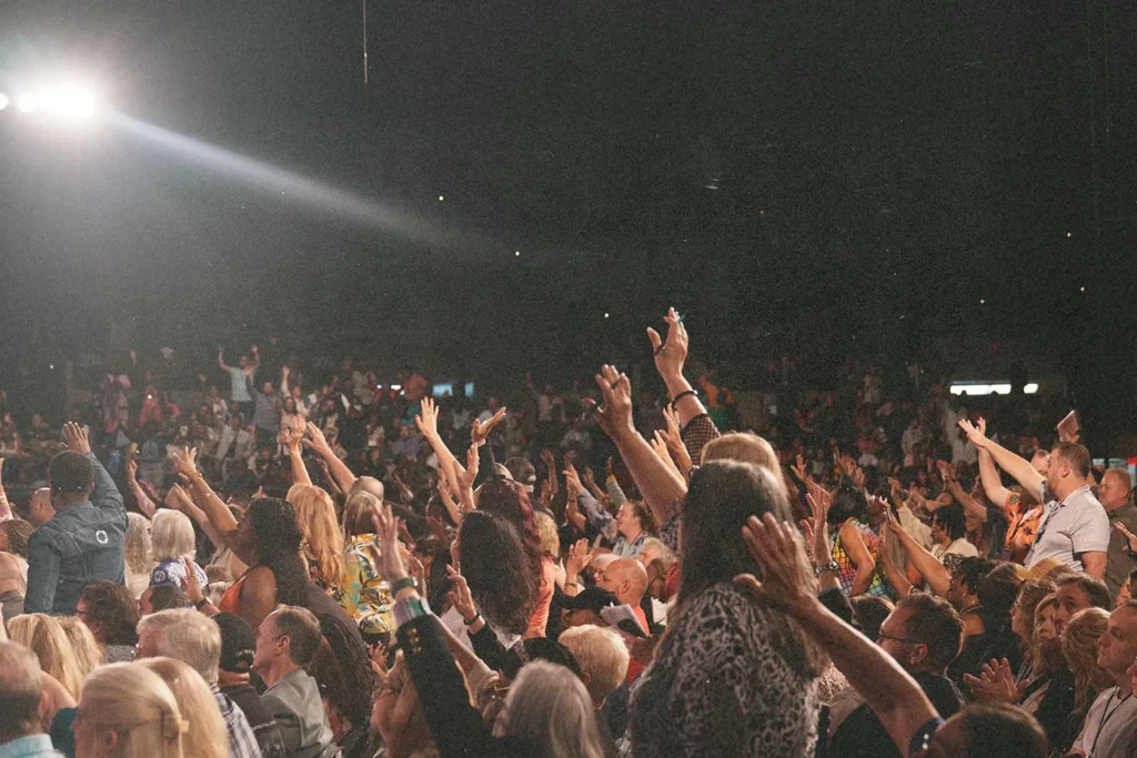 A large crowd gathers in an auditorium with many people raising their hands toward the stage under bright overhead lighting