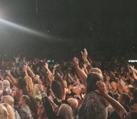 A large crowd gathers in an auditorium with many people raising their hands toward the stage under bright overhead lighting