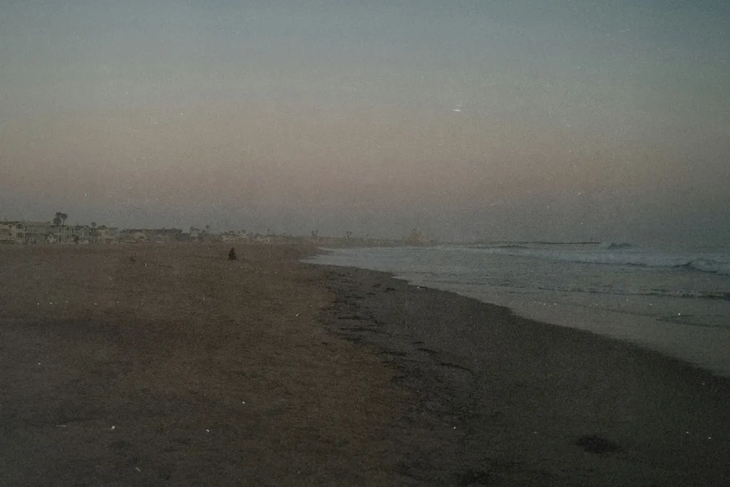 Wide, empty beach at dusk with gentle waves rolling onto the shore and faint silhouettes of distant buildings along the coastline