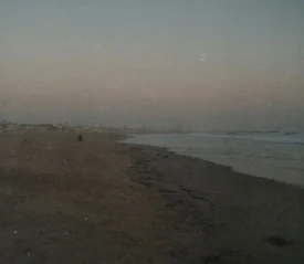 Wide, empty beach at dusk with gentle waves rolling onto the shore and faint silhouettes of distant buildings along the coastline