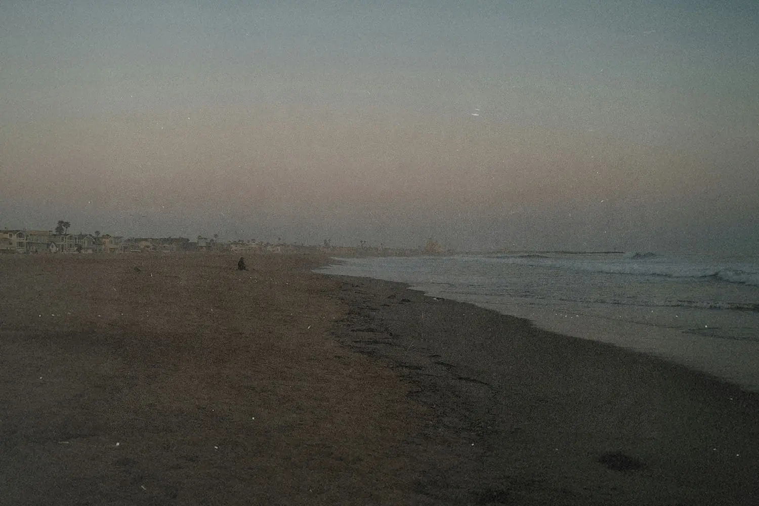 Wide, empty beach at dusk with gentle waves rolling onto the shore and faint silhouettes of distant buildings along the coastline