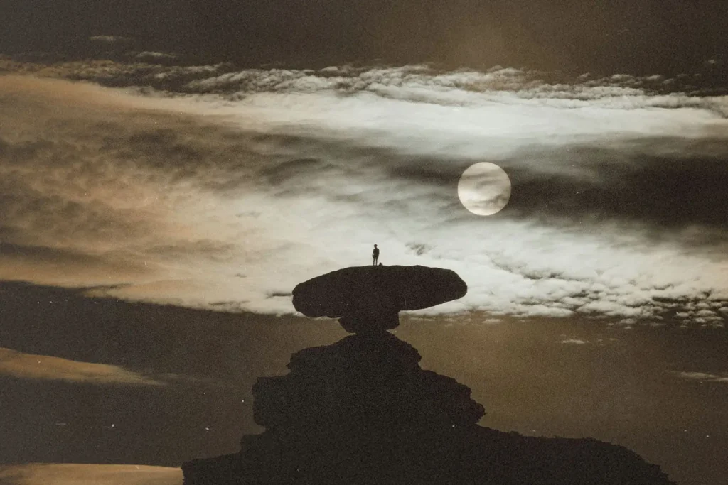 A lone figure stands on top of a large, flat rock formation beneath dramatic clouds and a bright, glowing moon