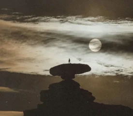 A lone figure stands on top of a large, flat rock formation beneath dramatic clouds and a bright, glowing moon