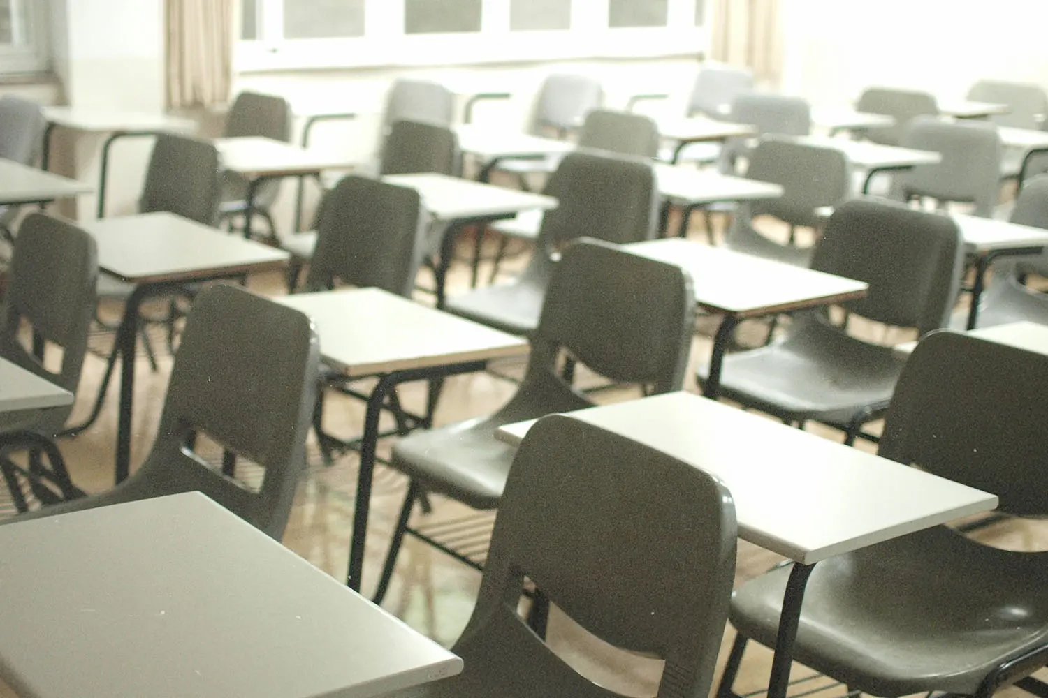 Rows of empty desks and chairs fill a bright classroom, with sunlight coming through large windows at the back of the room