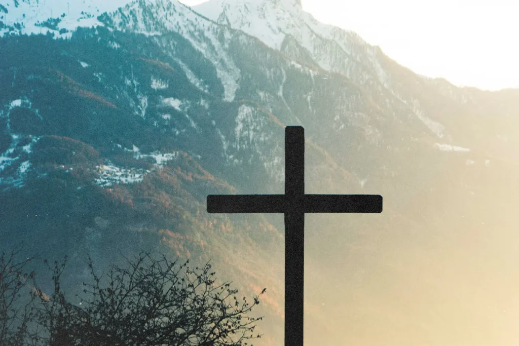 Silhouette of a cross set against sunlit mountains and a soft morning sky
