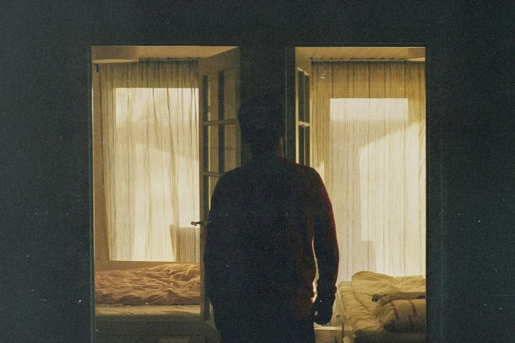 Silhouetted figure standing in a dimly lit doorway between two softly lit bedrooms with sheer curtains
