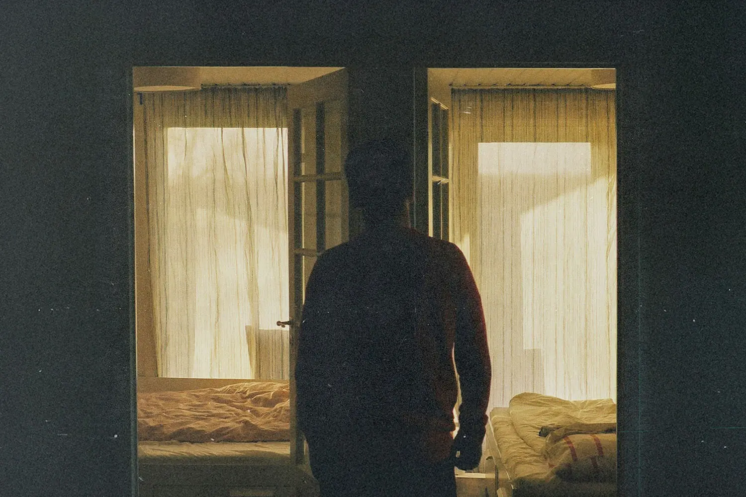 Silhouetted figure standing in a dimly lit doorway between two softly lit bedrooms with sheer curtains