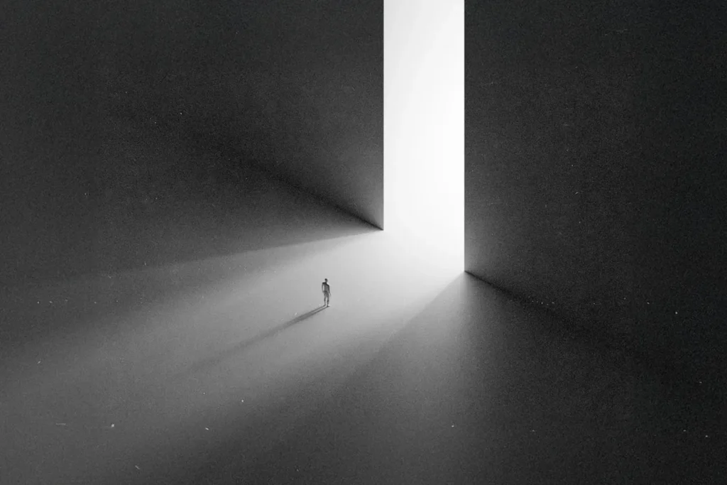 A small figure stands alone in a vast dark space, walking toward a towering doorway filled with bright white light