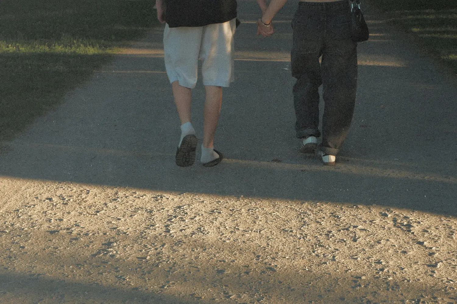 Two people walking hand in hand down a sunlit pathway in a park.