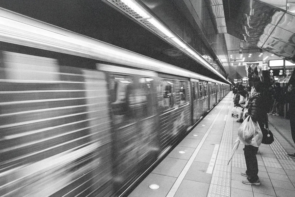 Subway train speeding past a platform, motion-blurred as it arrives or departs, while commuters stand waiting along the edge under bright overhead lights.