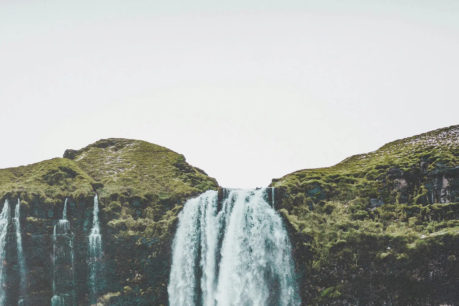 Wide waterfall cascading over a grassy cliff into a rocky valley under a bright, overcast sky