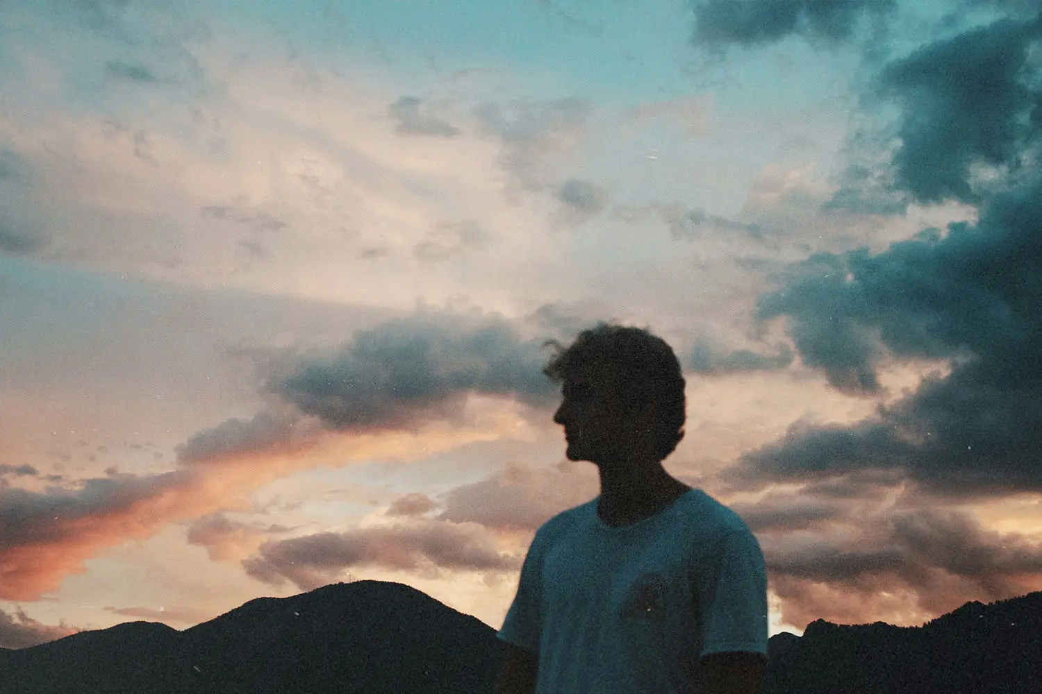 A person stands outdoors at sunset with mountains silhouetted behind them and colorful clouds filling the sky