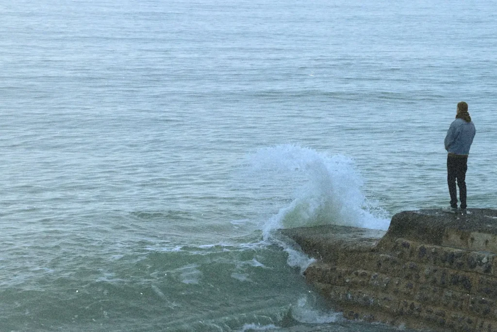 A person stands near the edge of a concrete ledge as a wave crashes against it, sending water upward