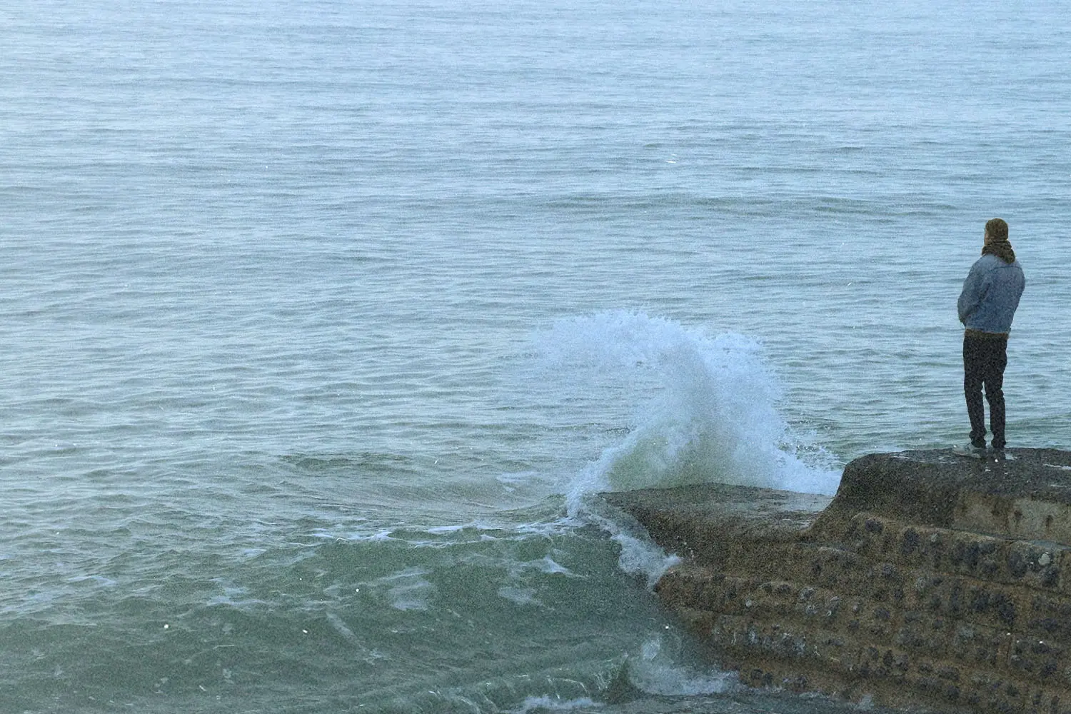 A person stands near the edge of a concrete ledge as a wave crashes against it, sending water upward