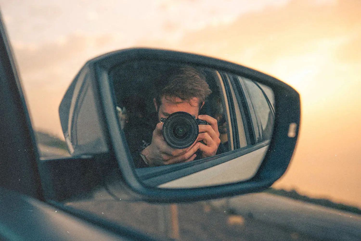 Photographer holding a camera reflected in a car’s side‑view mirror during a warm sunset drive