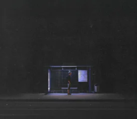 Single person stands under the dim blue light of a bus stop shelter at night, surrounded by darkness