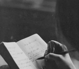 A person writes in a journal with a pencil, the open notebook filled with handwritten notes in soft, natural light