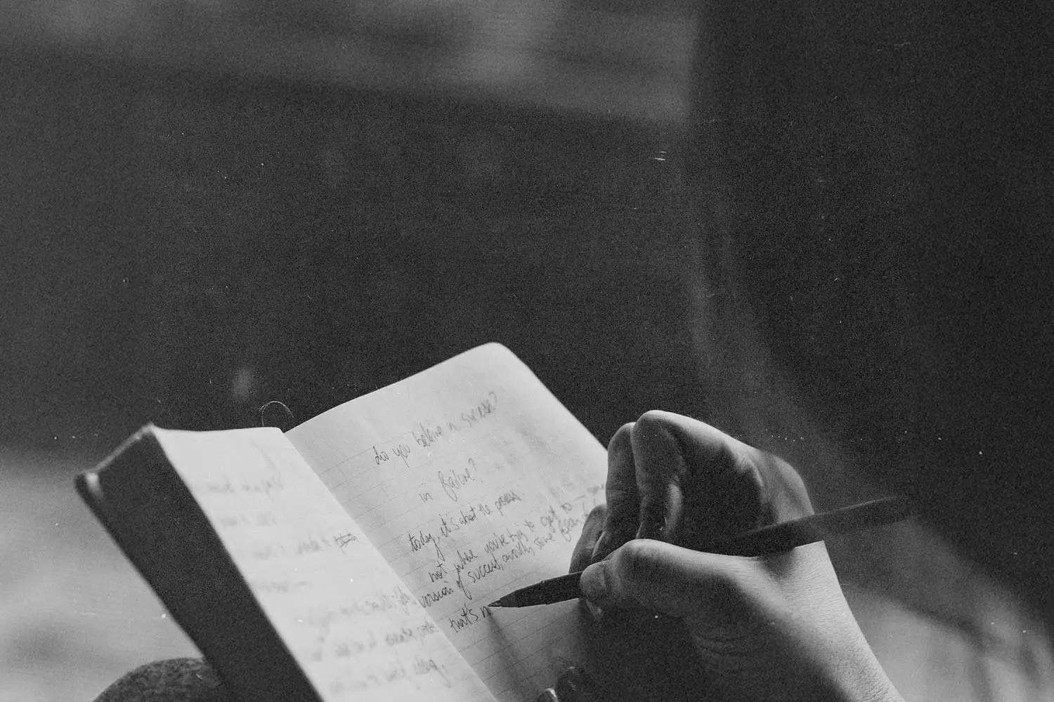 A person writes in a journal with a pencil, the open notebook filled with handwritten notes in soft, natural light