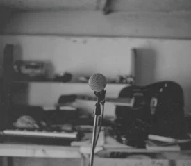 Single microphone on a stand in the center of a dimly lit room filled with musical instruments and equipment