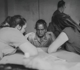 Three people sitting around a table in a quiet room, leaning in together in a moment of prayer