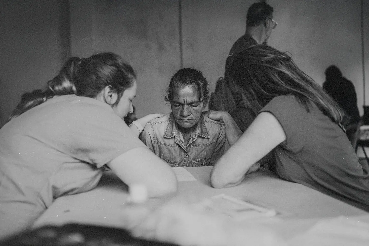 Three people sitting around a table in a quiet room, leaning in together in a moment of prayer