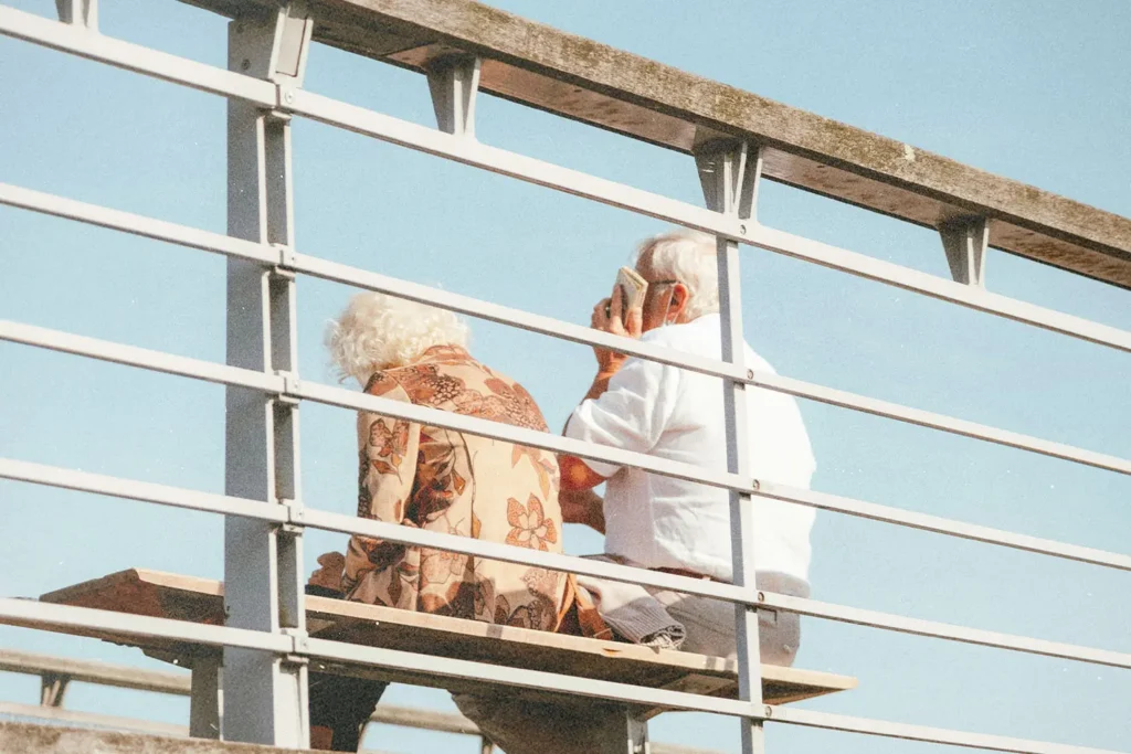 Two people sit on a bench outdoors behind a metal railing, with one holding a phone and clear blue sky in the background