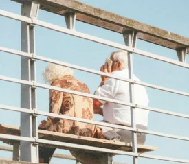 Two people sit on a bench outdoors behind a metal railing, with one holding a phone and clear blue sky in the background