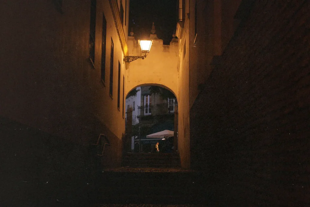 Narrow dimly lit alley leading to an archway with a warm lantern glowing above it at night