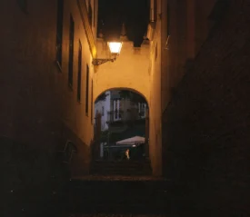 Narrow dimly lit alley leading to an archway with a warm lantern glowing above it at night