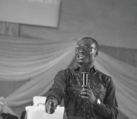 Speaker holding a microphone and gesturing toward the audience during an indoor event with draped fabric in the background