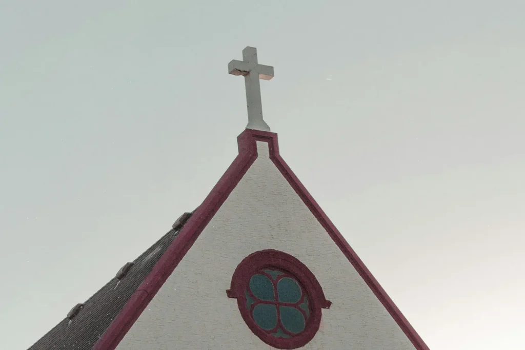 Steeple of a small church with a white cross on top and a circular stained‑glass window, set against a pale sky