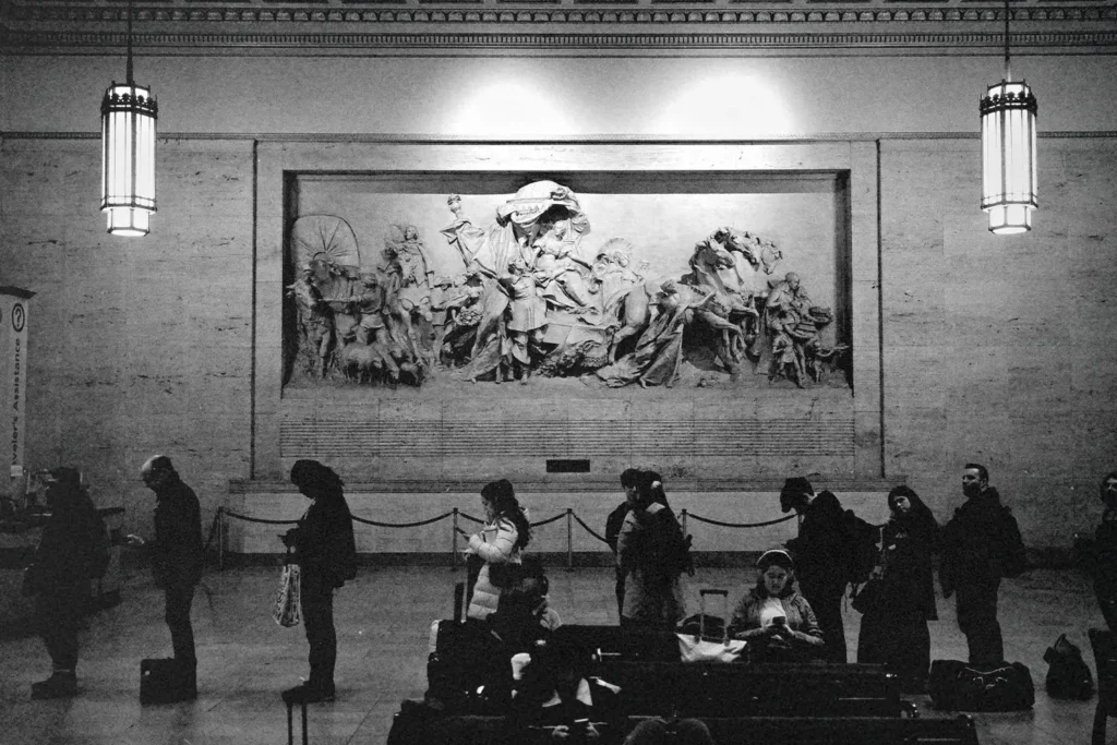 People standing and waiting inside a large hall with a detailed wall sculpture depicting historical figures, lit by tall hanging lamps