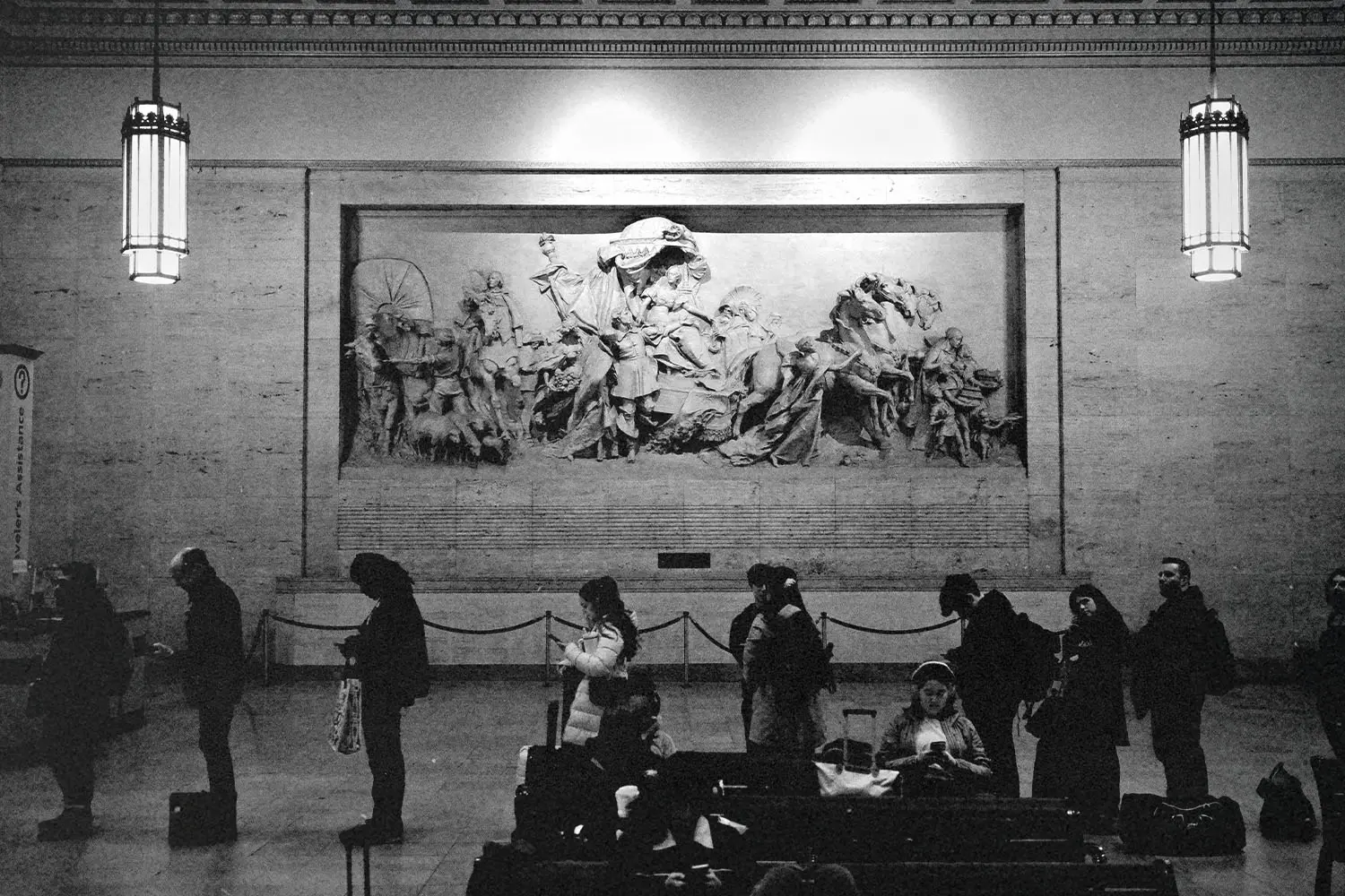 People standing and waiting inside a large hall with a detailed wall sculpture depicting historical figures, lit by tall hanging lamps