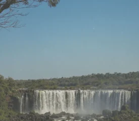 Wide waterfall cascading over a rocky cliff surrounded by lush green forest under a bright blue sky