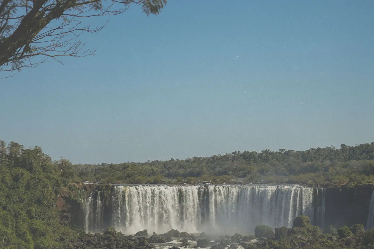 Wide waterfall cascading over a rocky cliff surrounded by lush green forest under a bright blue sky