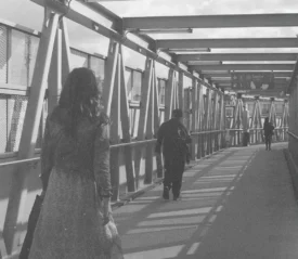 People walking through a long, covered pedestrian walkway with metal beams casting shadows along the path