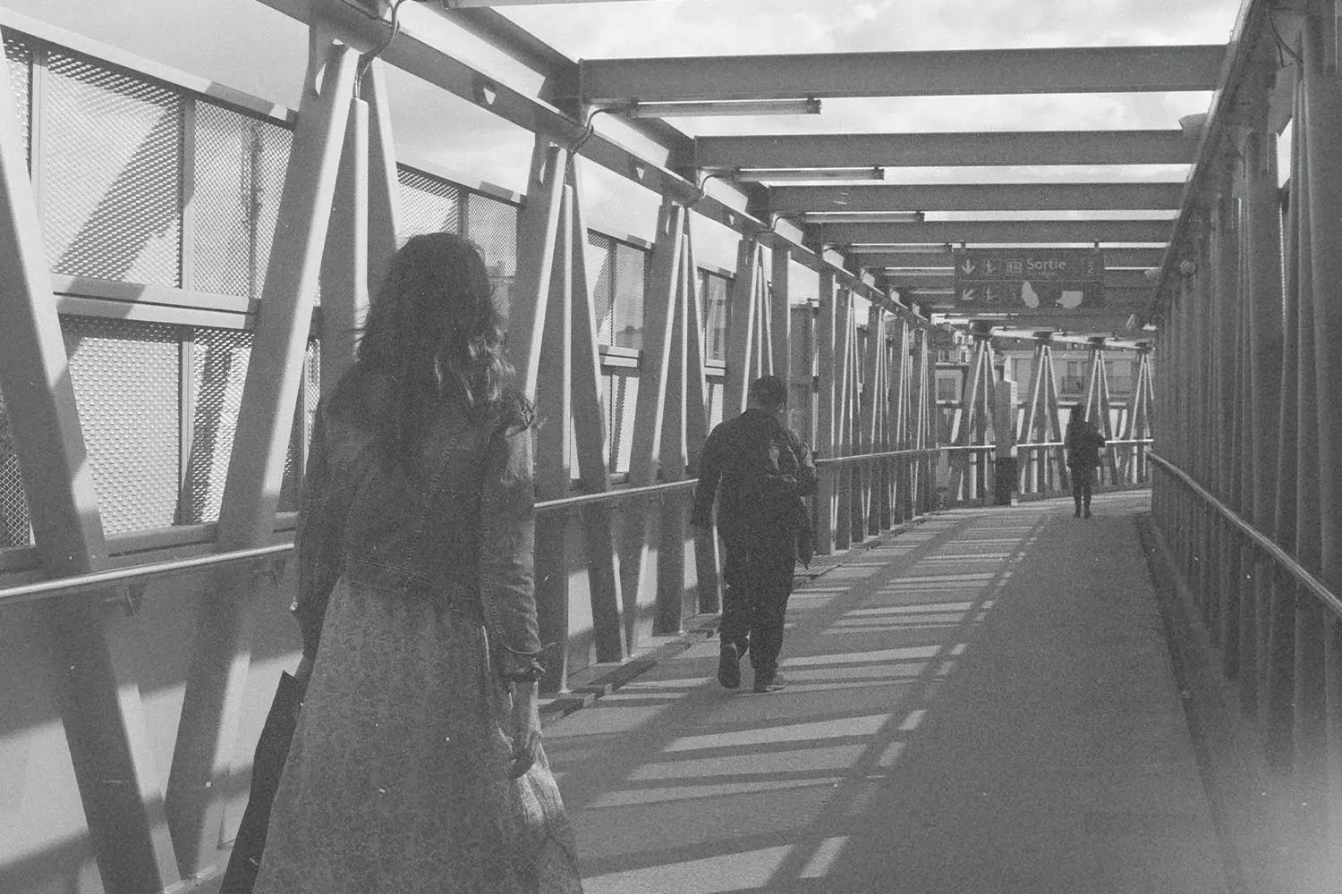 People walking through a long, covered pedestrian walkway with metal beams casting shadows along the path