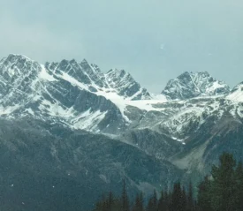 Snow‑covered mountain peaks rising sharply above dark forested slopes under a pale, overcast sky