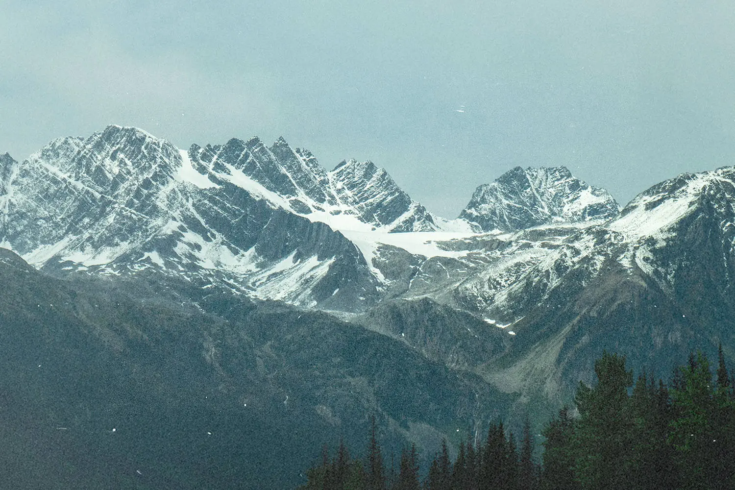 Snow‑covered mountain peaks rising sharply above dark forested slopes under a pale, overcast sky