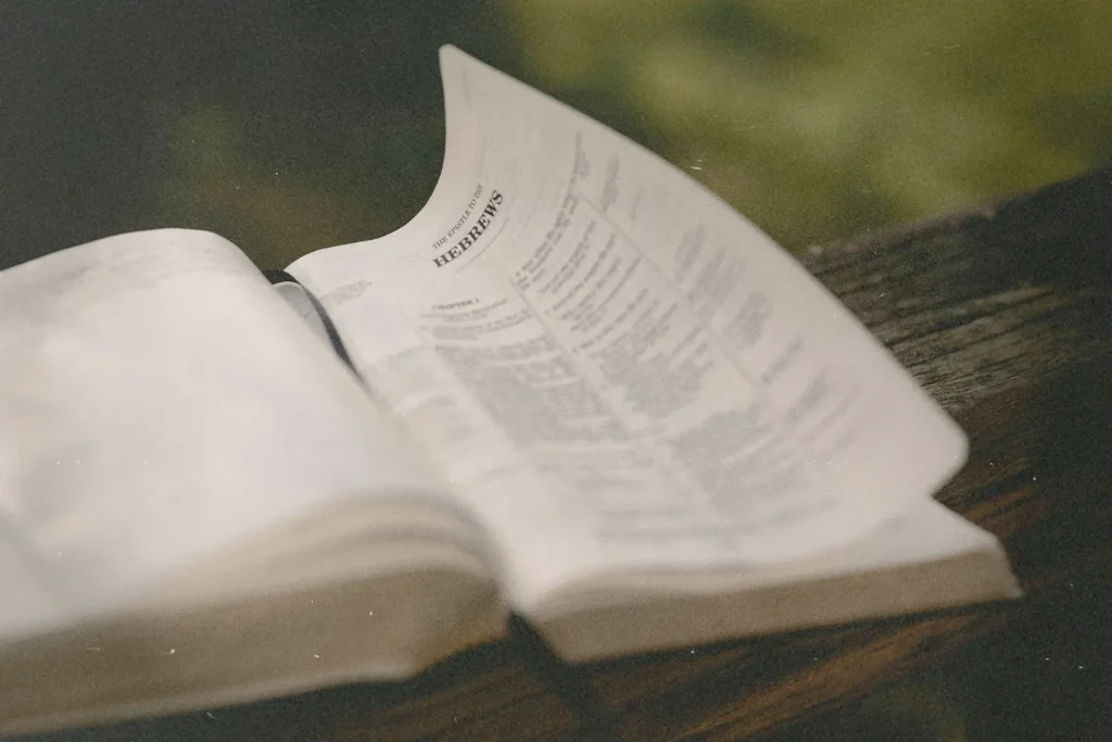 Open Bible on a wooden surface with a page from Hebrews gently lifting in the breeze outdoors