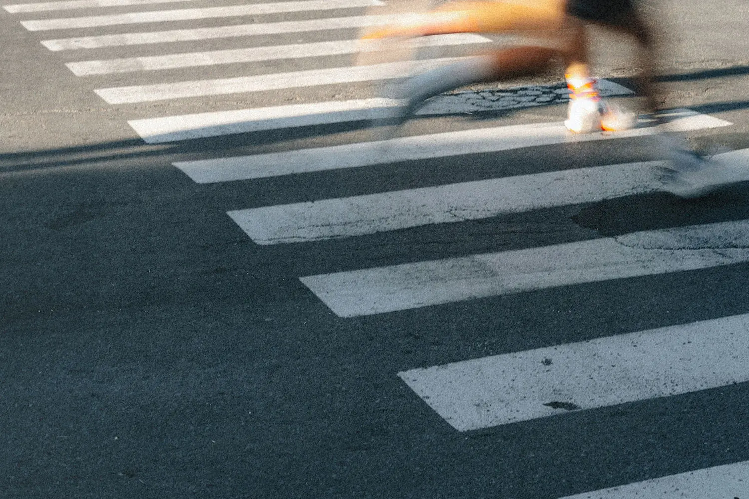 A blurred figure runs across a crosswalk, leaving a streak of motion over the white painted stripes on the pavement