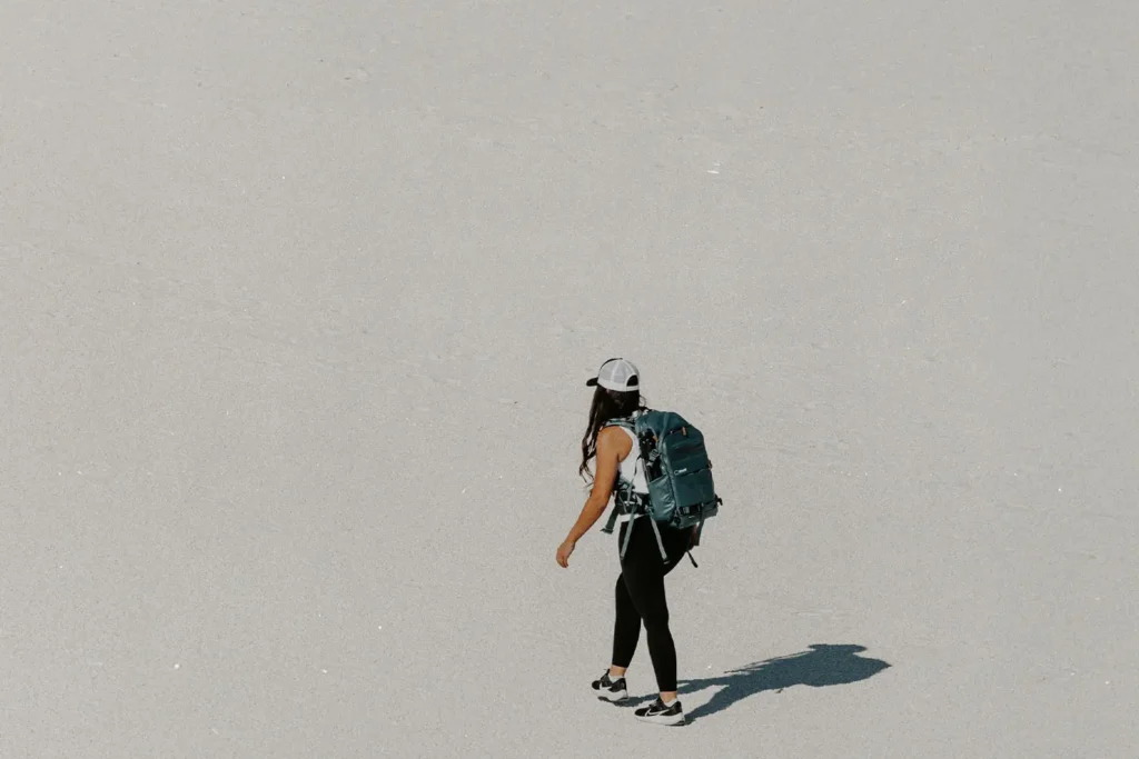 Person wearing a backpack walking alone across a wide, bright, sandy landscape, casting a long shadow in the sun
