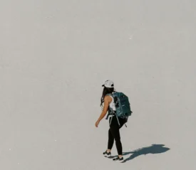 Person wearing a backpack walking alone across a wide, bright, sandy landscape, casting a long shadow in the sun