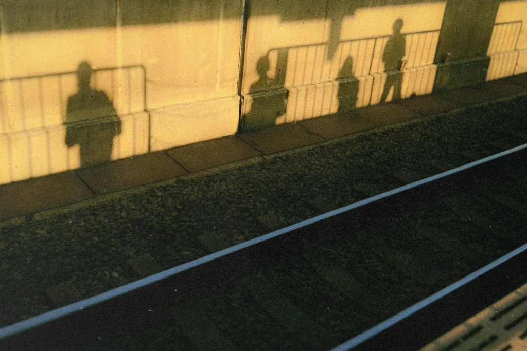 Long shadows of people standing on a train platform stretch across the wall and onto the ground in warm golden sunlight