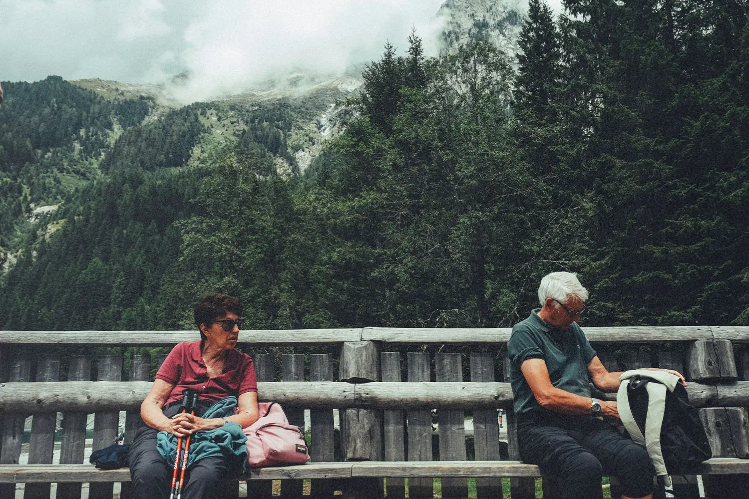 Two people sitting on a long wooden bench in a mountainous forest setting, with one person holding hiking poles and the other looking into a backpack